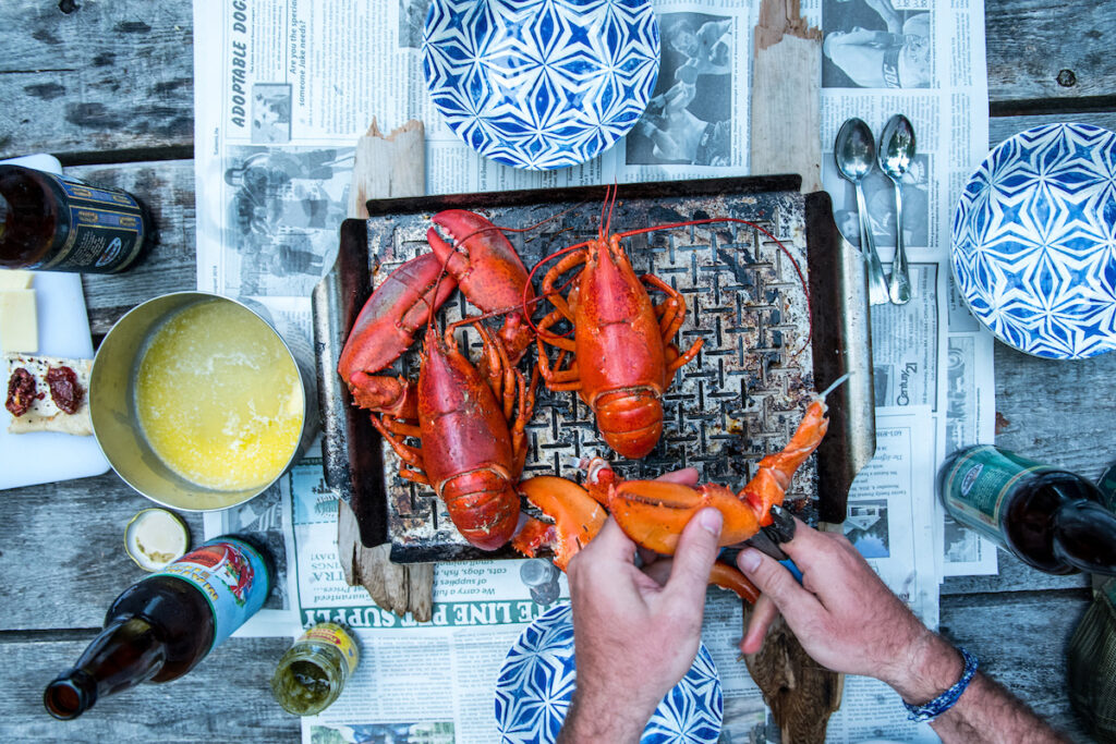 Two cooked lobsters on a tray, surrounded by blue and white plates, a bowl of melted butter, beer bottles, and utensils on a newspaper-covered wooden table. Hands are cracking a lobster claw at a summer festival in Maine