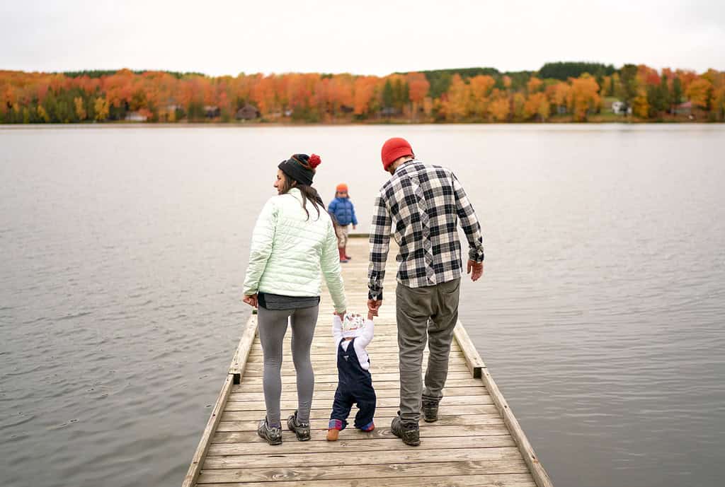 A family walks on a wooden dock by a lake with autumn trees in the background.