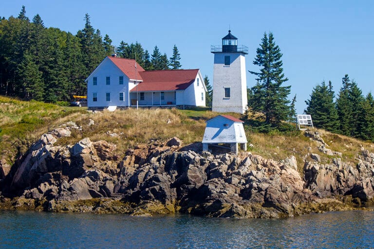 Lighthouse and white house with red roof on a rocky shoreline surrounded by trees and water on a clear day.