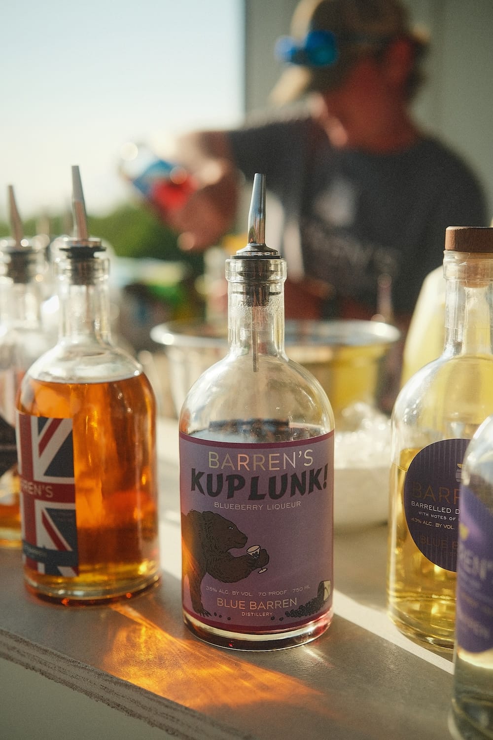 A bottle of Blue Barren’s Kuplunk blueberry liqueur with a label featuring a bear, surrounded by other glass bottles on a bar.