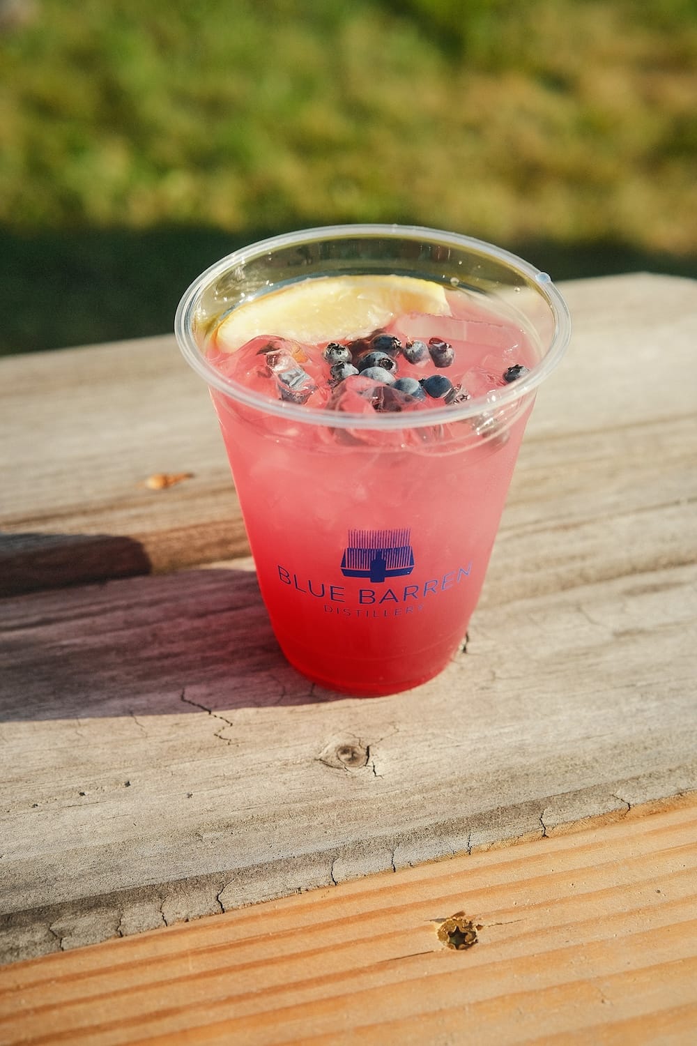 A pink cocktail in a clear plastic cup with the Blue Barren Distillery logo, garnished with blueberries and a lemon slice, sitting on a wooden surface.