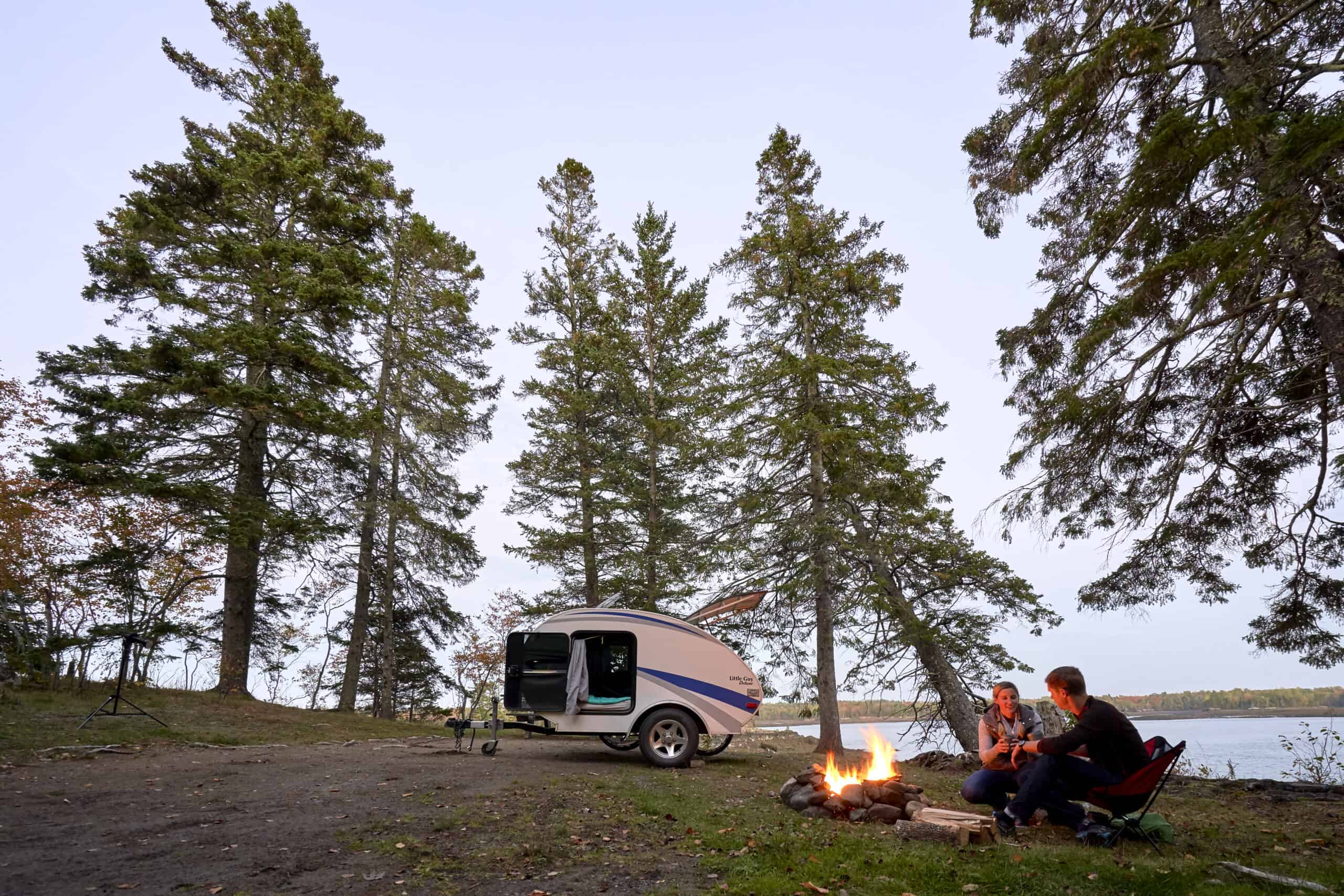 A couple sitting by a campfire with a mini-camper trailer in the back ground
