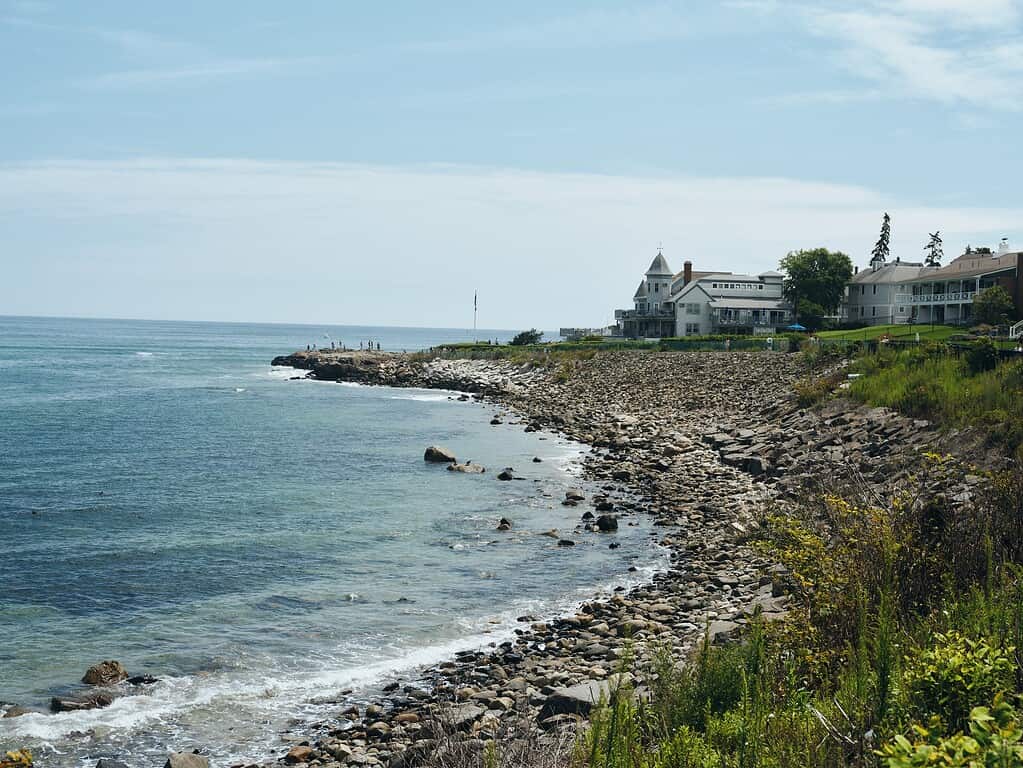 Rocky shoreline with gentle waves, coastal vegetation, and several houses overlooking the ocean under a partly cloudy sky.