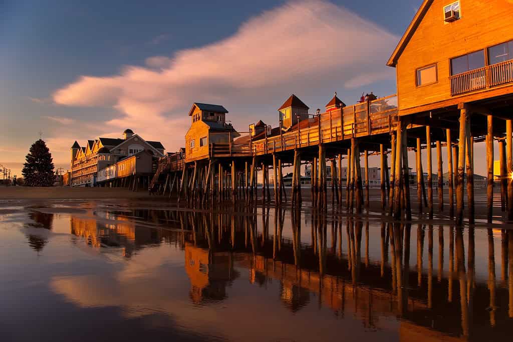 Wooden pier with buildings extends over calm reflective water at sunset, under a sky with scattered clouds.