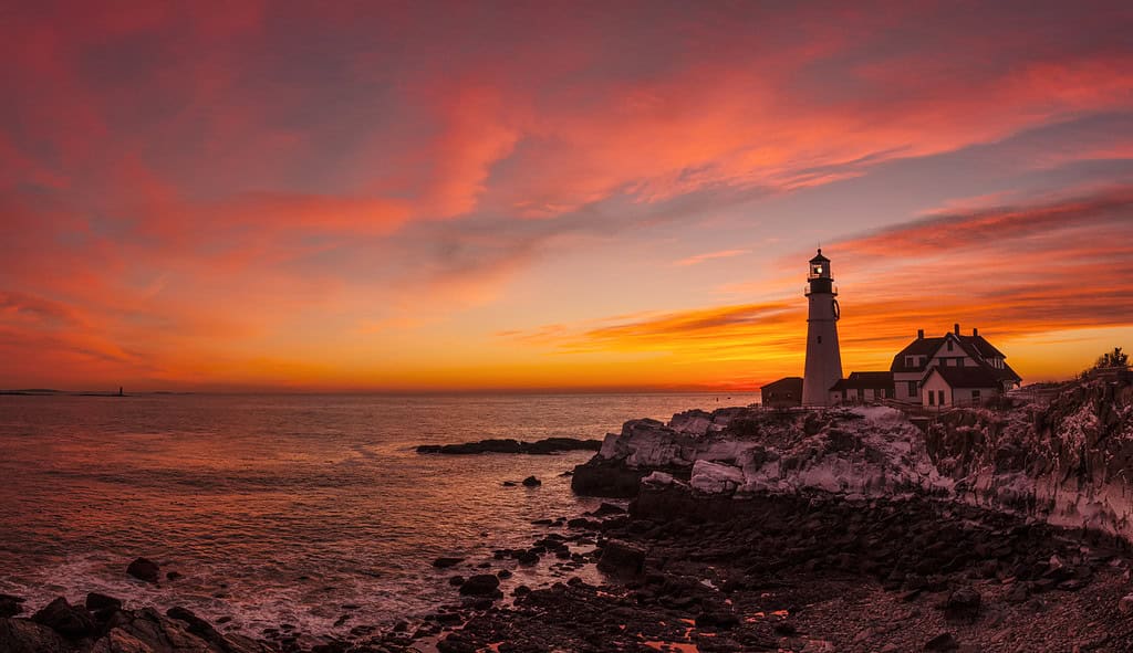 A lighthouse stands on a rocky coastline during a vibrant orange and pink sunset.
