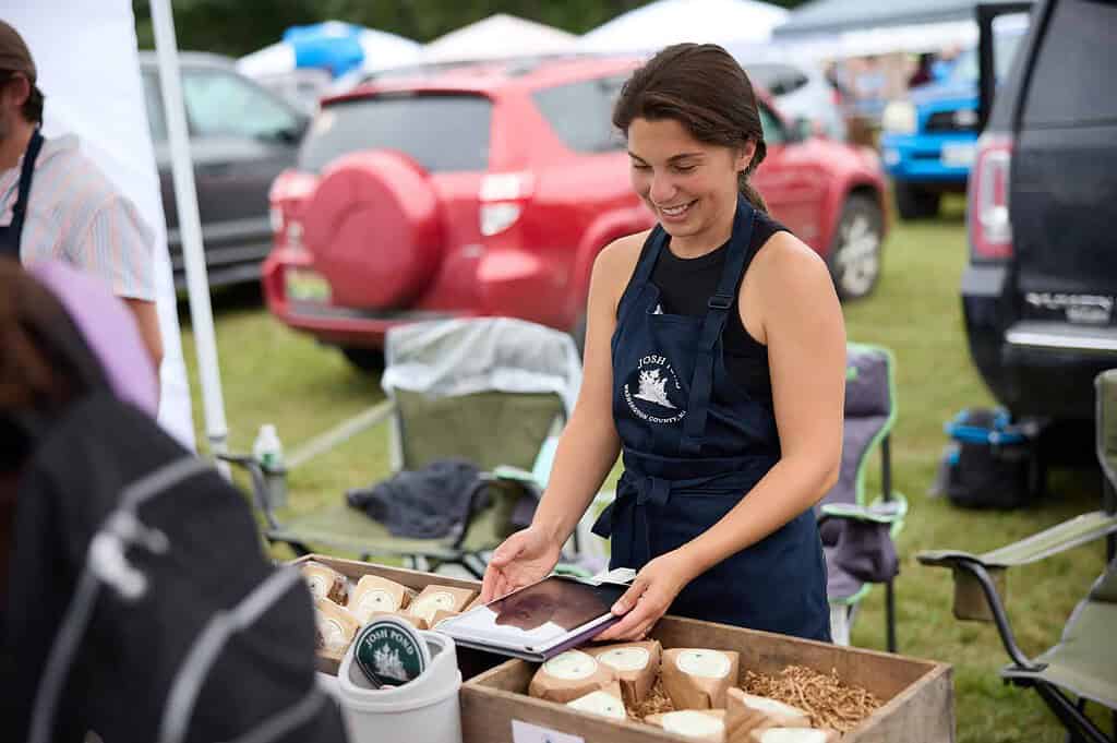 A smiling vendor in a blue apron holds a tablet at an outdoor market stall featuring packaged goods.