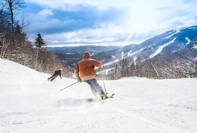 Two skiers descend a snowy mountain slope surrounded by trees under a bright sky.
