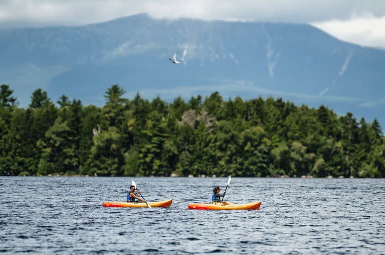Two people kayaking on a lake with a distant mountain and dense forest backdrop under a cloudy sky.