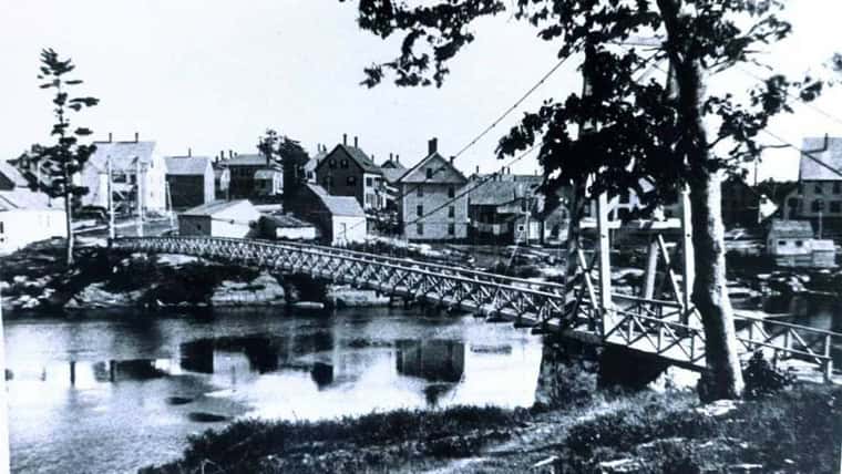 Black and white photo of a small town with a narrow bridge crossing over a river. Homes and trees line the riverbank, reflecting in the water. Some utility poles and wires are visible.