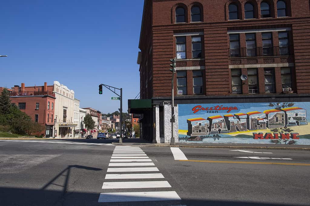 Street view featuring a "Greetings from Bangor, Maine" mural on a brick building with downtown shops and a clear blue sky.