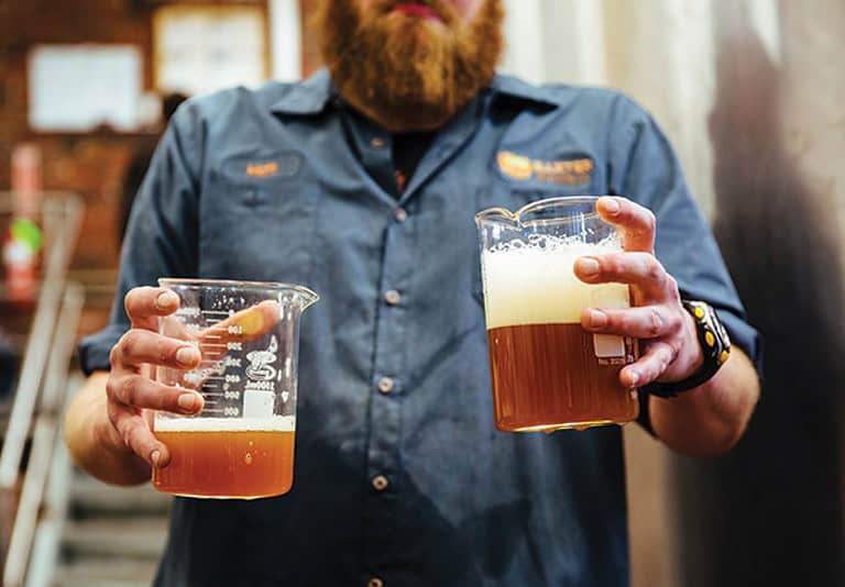A person with a beard and blue shirt holds two beakers of beer inside a brewery.