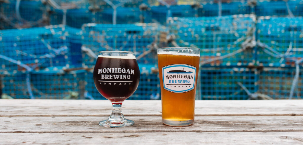 Two glasses of beer from Monhegan Brewing Company on a wooden table, with stacks of blue fishing traps in the background.