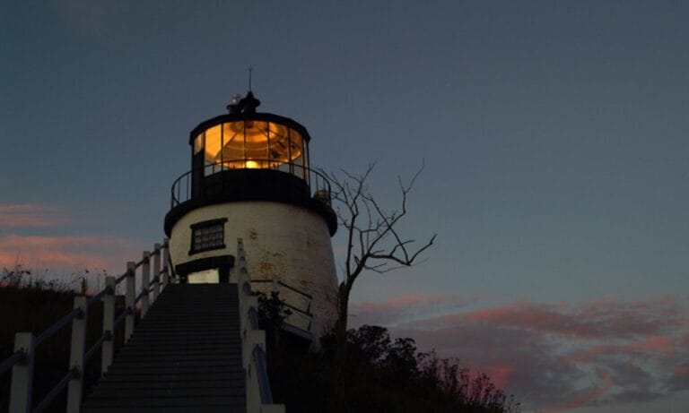 A lighthouse with its light on at dusk, viewed from the base of the stairs leading up to it. Trees and bushes are silhouetted against a darkening sky with tinges of pink clouds, creating a serene backdrop for Owl's Head Lighthouse.