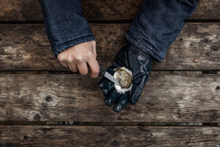 A person wearing a black glove uses a knife to open an oyster on a weathered wooden surface.
