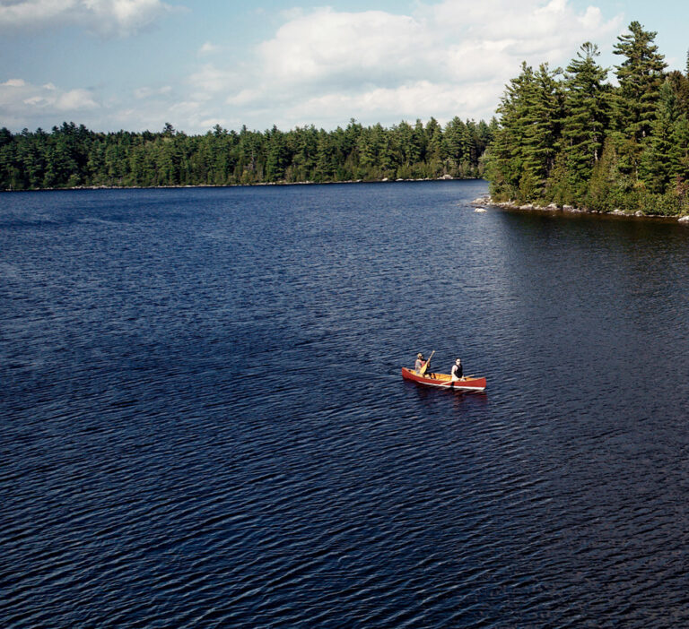 Two individuals paddle a red canoe on a calm lake surrounded by dense forest under a blue sky with scattered clouds.