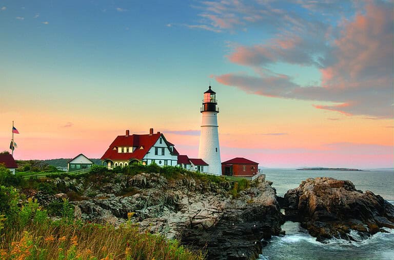 The Portland Head Lighthouse and adjoining buildings with red roofs stand on a rocky coastline under a colorful sunset sky. An American flag is visible near the buildings.