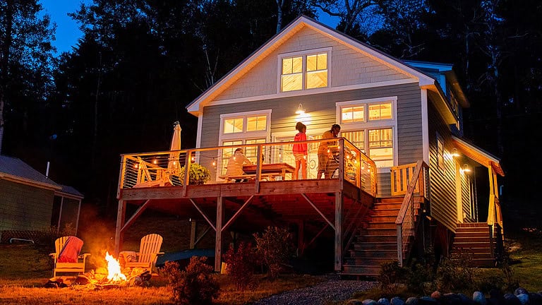 A two-story house with lit outdoor lights and a spacious deck at dusk. Two people stand on the deck. A fire pit and two Adirondack chairs are in the foreground.
