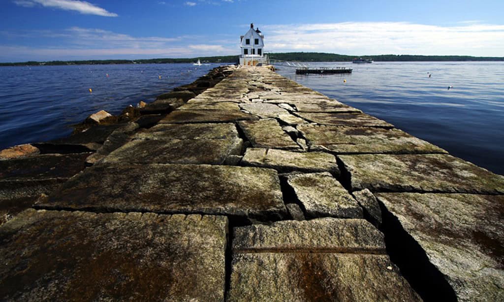 A walkway of large stones leads to the historic Rockland Breakwater Lighthouse, overlooking a calm body of water with a partly cloudy sky overhead.