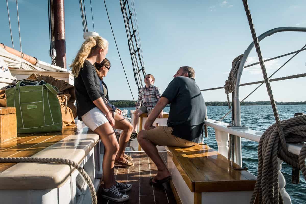 A group of people sitting and talking on a Windjammer Cruise in sunny weather, with a view of the water and distant shoreline.