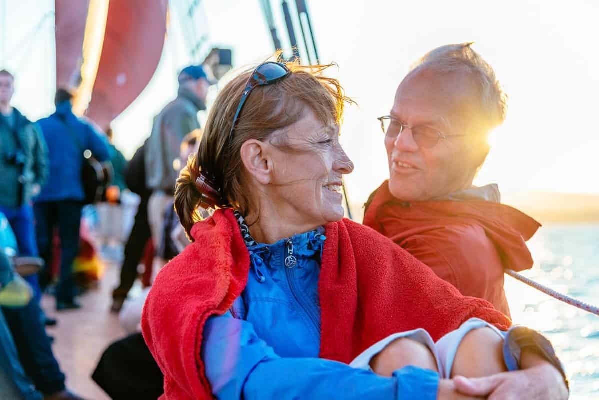 A woman and man smile at each other on a Windjammer Cruise during sunset. The woman is wrapped in a red blanket. Other people are in the background.