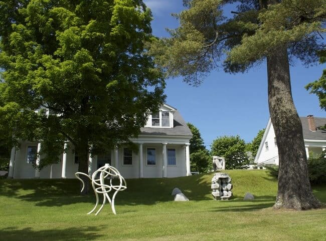 A white house with a porch is situated among green trees on a grassy lawn adorned with modern outdoor sculptures.