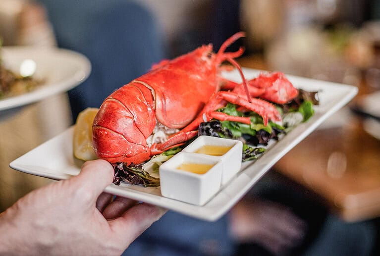 A whole cooked lobster served on a white plate with greens, lemon wedges, and two small containers of dipping sauce.