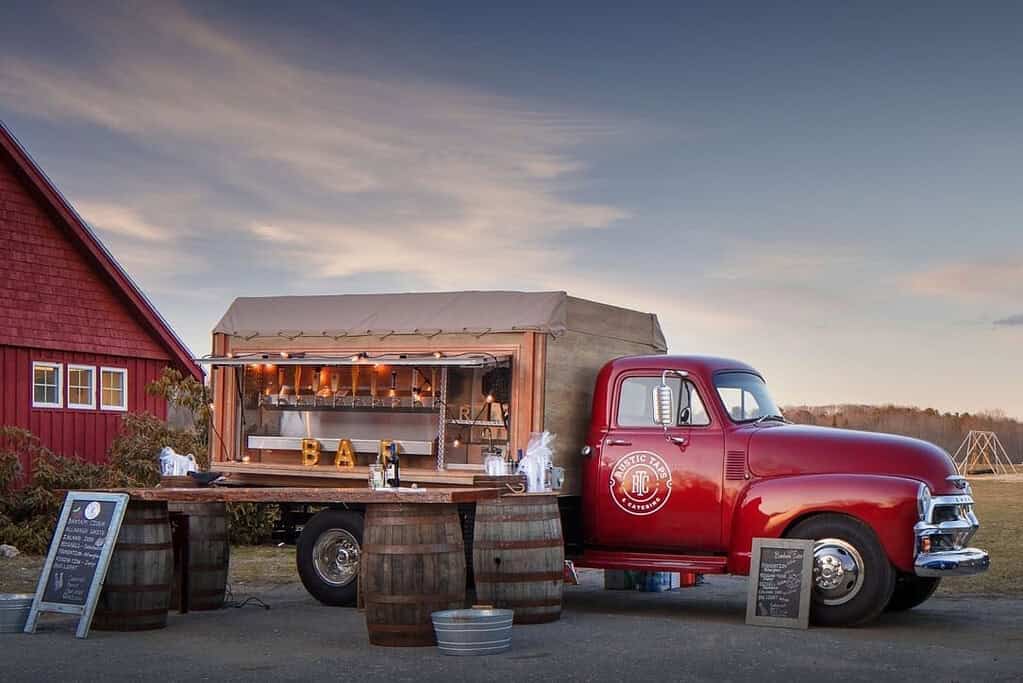 A vintage red truck converted into a bar with a wooden serving counter set on two barrels, parked near a red building. Signboards and metal tubs are placed around the setup in Maine.