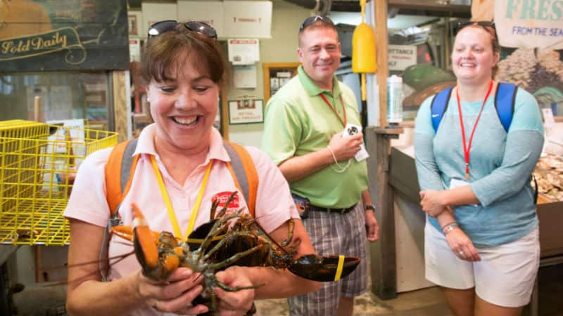 A woman holds a large lobster while two people stand and watch inside a seafood market.