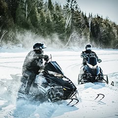 Two people wearing helmets riding snowmobiles on a snowy landscape with evergreen trees in the background.