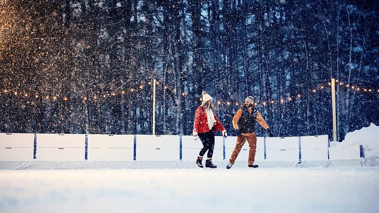 Two people ice skating on an outdoor rink, surrounded by snow-covered trees and illuminated by string lights in the evening.