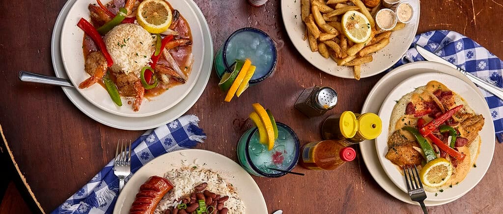 Three plates with various meals, including rice, shrimp, sausage, and fries, alongside two drinks with lemon slices, on a wooden table with a blue-checkered napkin.