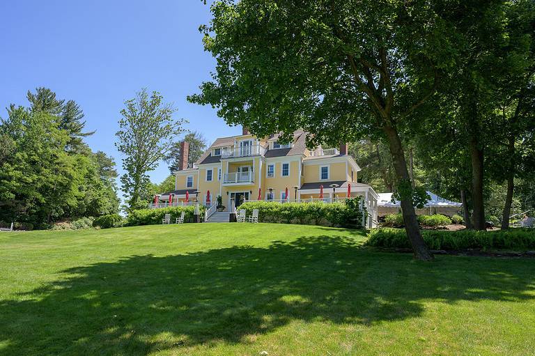 A large yellow house with multiple dormers and chimneys is set on a lush green lawn, surrounded by trees under a clear blue sky.