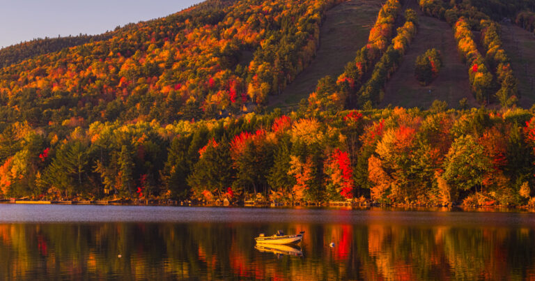 A small boat floats on a lake reflecting colorful autumn trees and a sunlit hillside.