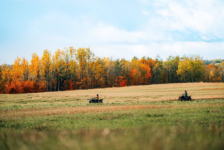 Two people ride ATVs across a grassy field with trees in the background, showing autumn colors in orange and yellow.