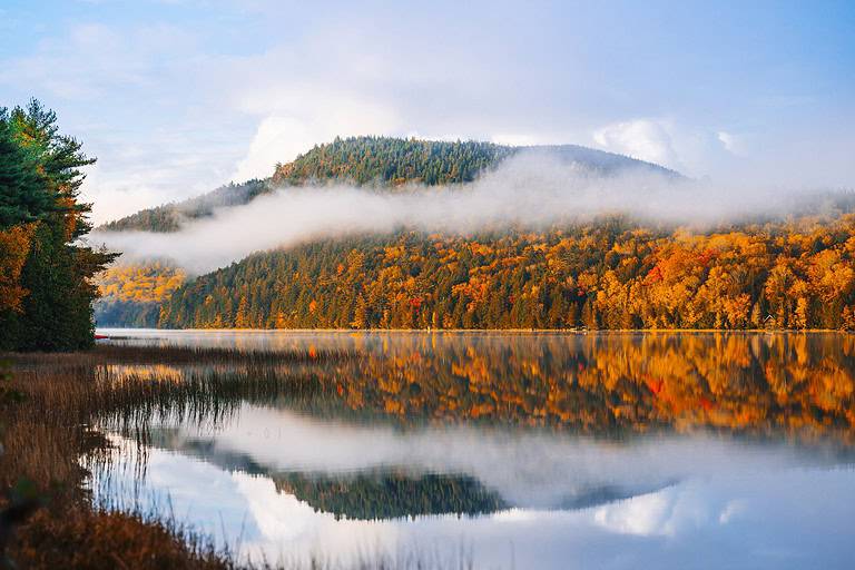 A calm lake reflects autumn-colored trees and a mist-covered hill, under a partly cloudy sky.