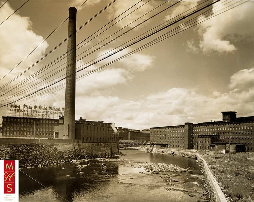 Historic mill buildings and smokestack along a river, with power lines crossing the sky.