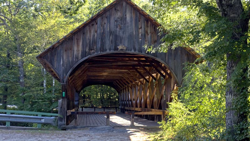 A rustic wooden covered bridge surrounded by trees and sunlight. The entrance is shadowed, and there are guardrails to the side.