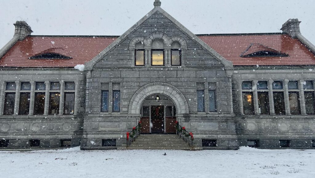 A historic stone building with red roofs, covered in snow. Snow falls in the foreground, and there are wreaths on the entrance.
