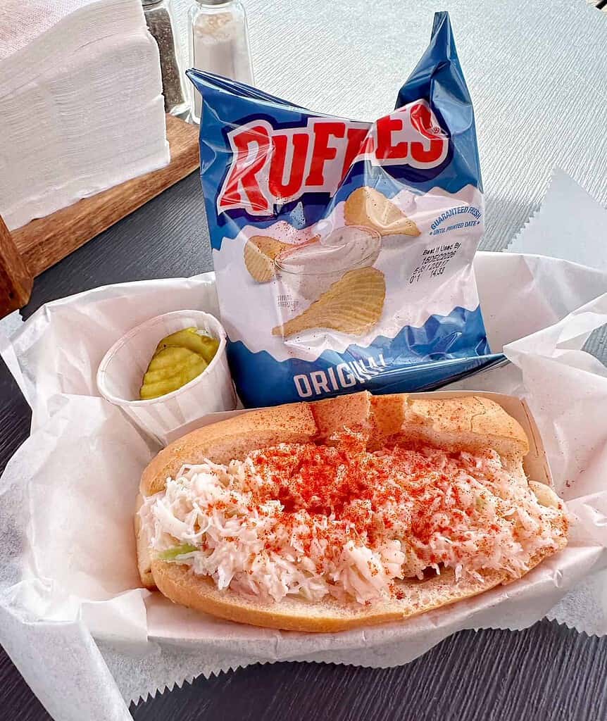 A lobster roll sprinkled with paprika in a paper-lined basket, served with a small cup of pickles and a bag of Ruffles potato chips.