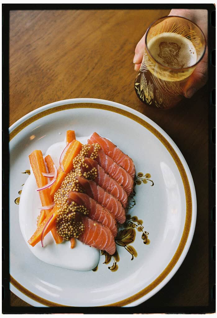 A plate with sliced cured salmon, pickled carrots, mustard seeds, and a white sauce next to a hand holding a glass of beer.