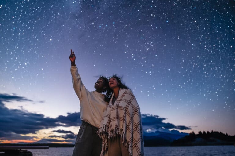 Two people stand outdoors under a star-filled night sky, one pointing upward, both smiling; mountains and water are visible in the background.