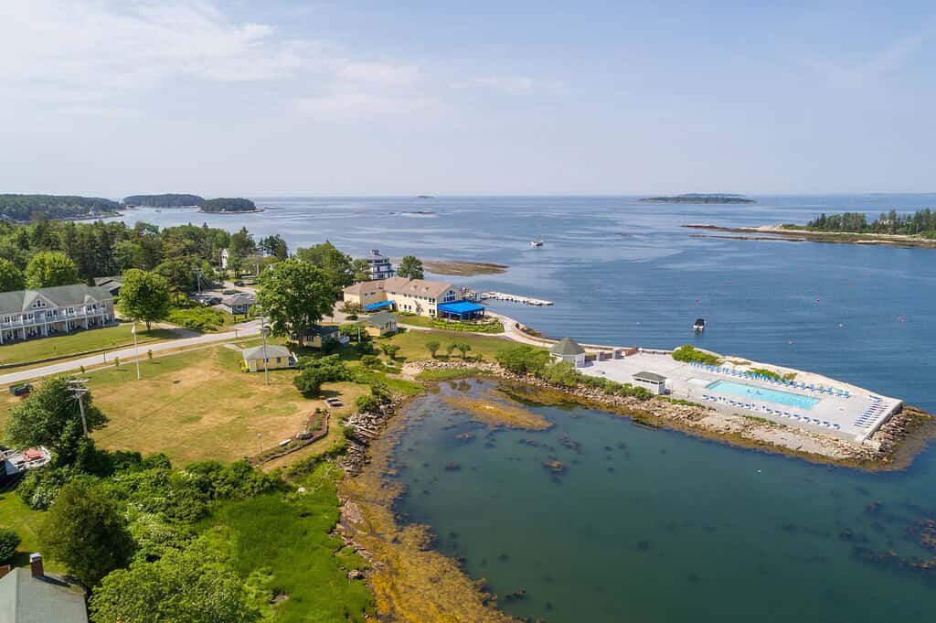 Aerial view of a coastal area with buildings, a swimming pool near the shoreline, grassy land, and calm blue water with small islands in the distance.