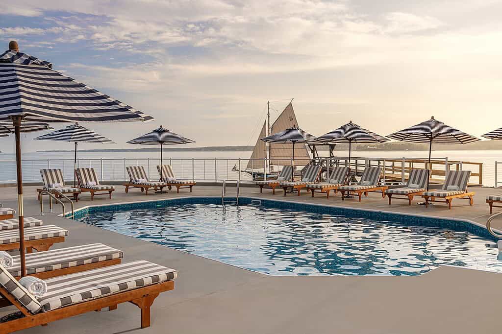 Outdoor pool area with striped lounge chairs and umbrellas overlooking the ocean; sailboat visible on the water in the background during sunset.