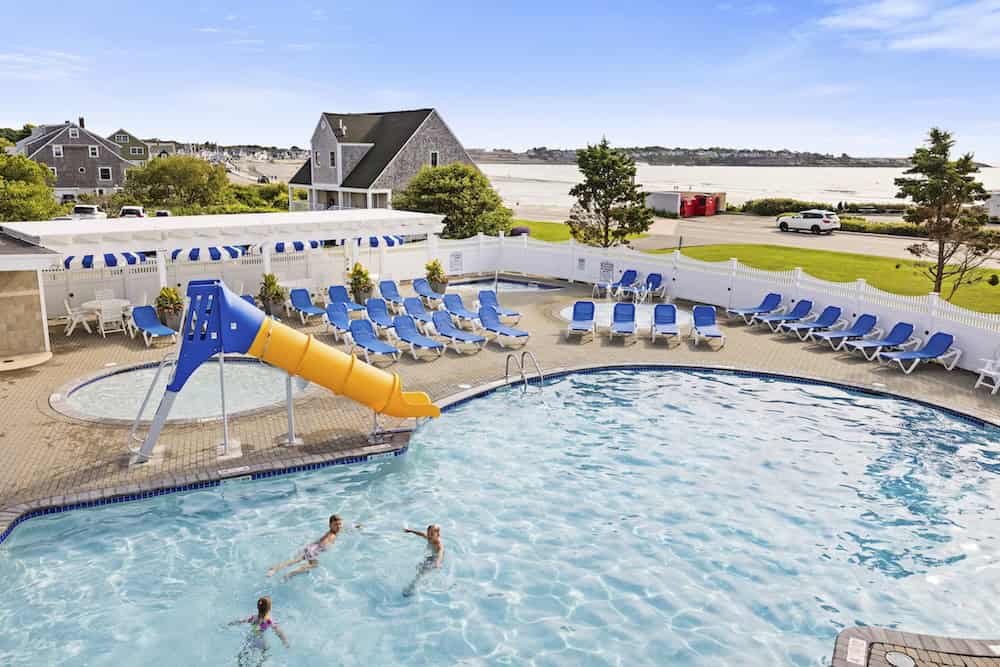 Outdoor swimming pool with a yellow slide, lounge chairs, and two people in the water; a small hot tub, grassy area, and houses are visible in the background under a clear sky.