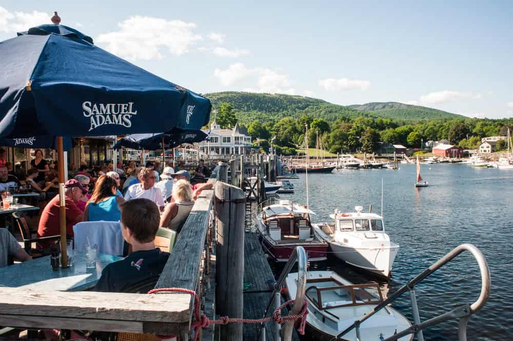 people sitting on a patio under sun umbrellas and overlooking the harbor with fishing boats docked