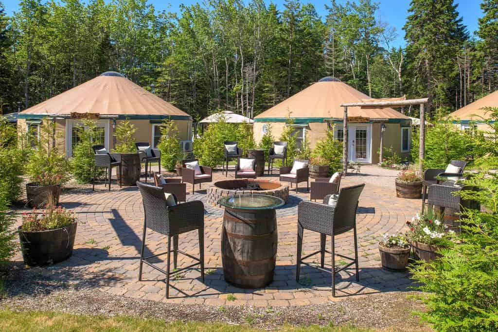 Circular outdoor seating area with wicker chairs and a firepit, surrounded by potted plants and three tan yurts in a wooded setting.