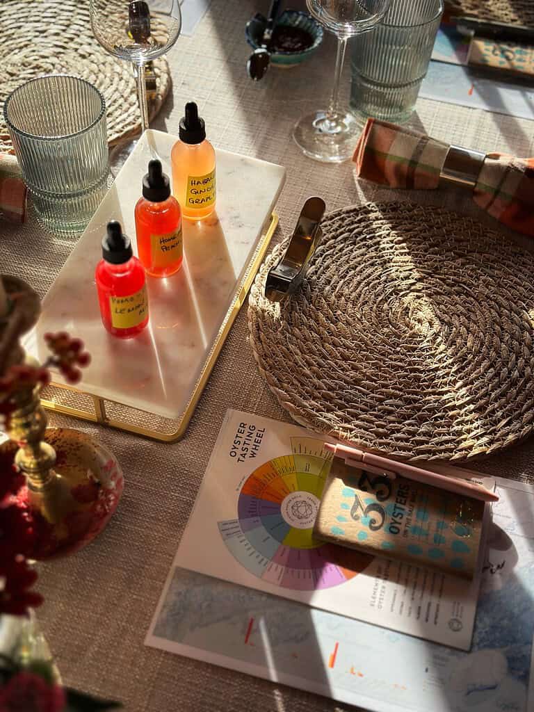 A tabletop display with three dropper bottles, a placemat, a glass, a wrapped chocolate bar, a color wheel chart, and wine glasses in warm sunlight.