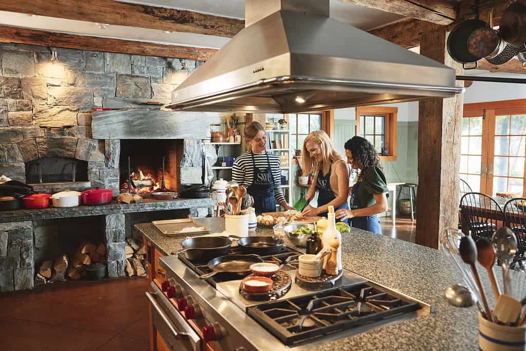 Three women prepare food together at a kitchen island in a rustic, well-lit kitchen with exposed beams and a stone fireplace.