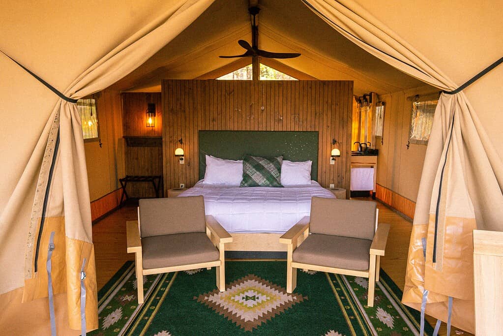 View of a glamping tent interior with a bed in the center, two armchairs, a patterned green rug, wooden walls, and visible lighting fixtures.