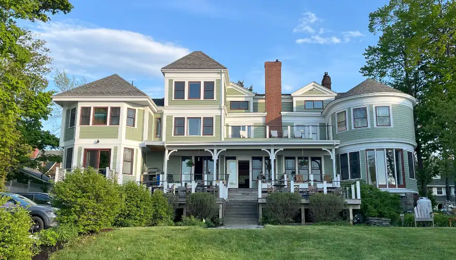 Large, light green Victorian-style house with a wraparound porch, multiple gables, bay windows, and a central brick chimney, set among trees and shrubs on a sunny day.